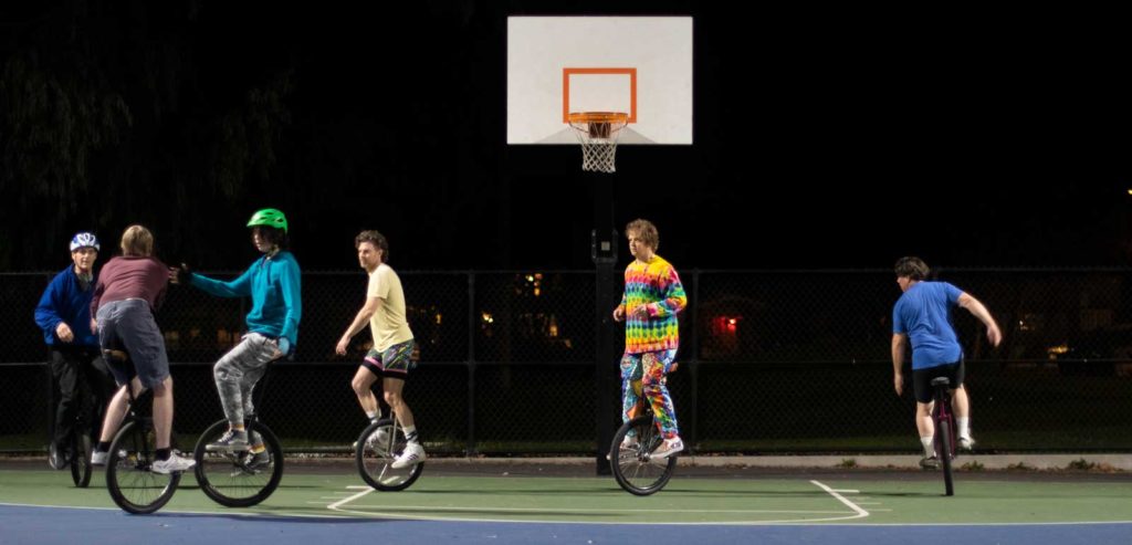 Six people on unicycles playing basketball on a lit court at night