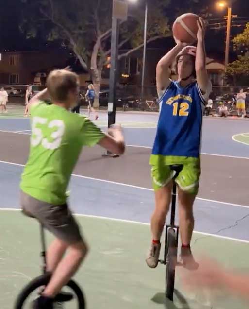 Two men on unicycles play basketball on a well-lit court at night. One is in the process of taking a shot, the other is rushing to defend. The one taking the shot is wearing a jersey that reads "REVOLUTION", with the number 12.
